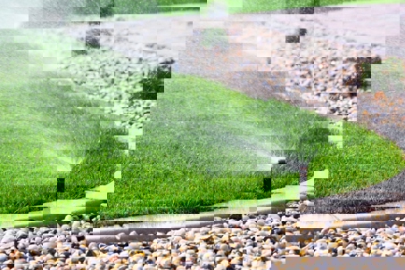 A lawn sprinkler waters a patch of healthy, bright green grass next to decorative rocks and a concrete curb.