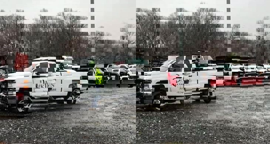 An outdoor photo shows a fleet of white pickup trucks with snow plows in a parking lot.