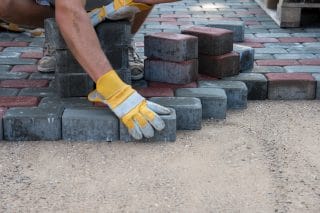A landscaper wearing work gloves lays pavers on a bed of sand