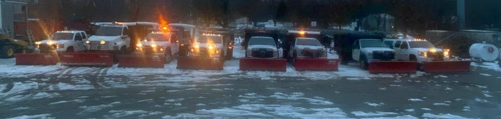 A row of snowplows, illuminated with orange lights, are parked in a snowy lot.