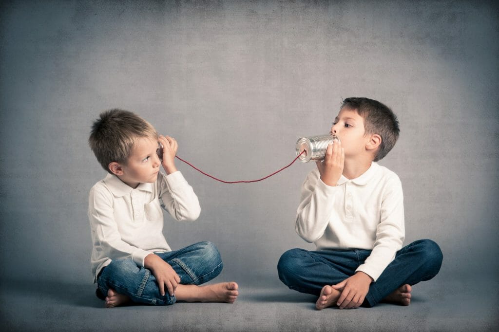 Two boys are sitting across from each other, communicating using a tin can telephone
